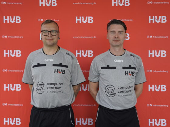 Zwei Männer in grauen Kempa-Uniformen stehen vor einem roten Hintergrund mit dem HVB-Logo. Beide blicken nach vorne, stehen nebeneinander und haben die Arme hinter dem Rücken verschränkt.