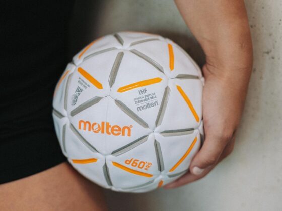 Eine Person hält einen weißen, geschmolzenen d60-Pro-Handball mit orangefarbenen und grauen Akzenten in der Hand und steht neben einer hellen Wand – ein ideales Bild, das das Engagement und die Präzision im Nachwuchsleistungssport widerspiegelt.