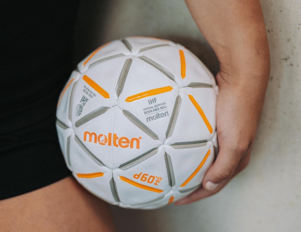 Eine Person hält einen weißen, geschmolzenen d60-Pro-Handball mit orangefarbenen und grauen Akzenten in der Hand und steht neben einer hellen Wand – ein ideales Bild, das das Engagement und die Präzision im Nachwuchsleistungssport widerspiegelt.