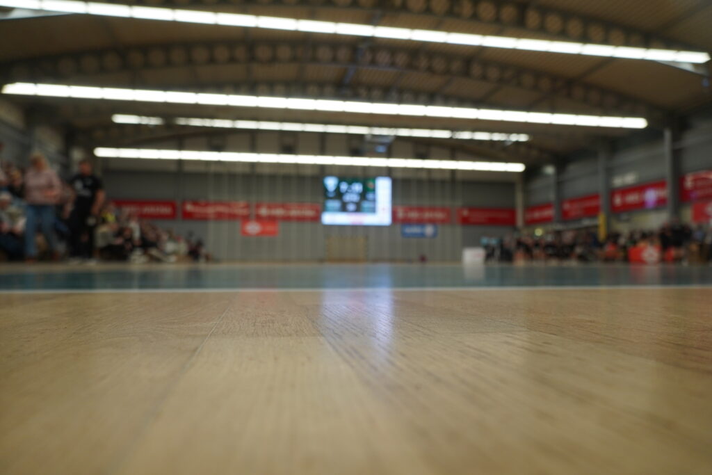Nahaufnahme eines Turnhallenbodens mit unscharfem Hintergrund, auf dem Menschen auf den Tribünen sitzen und eine große Anzeigetafel zu sehen ist – alles in einer gut beleuchteten Sporthalle: eine dynamische Kulisse, die häufig für den Nachwuchsleistungssport genutzt wird, in der junge Sportler trainieren und Wettkämpfe bestreiten.