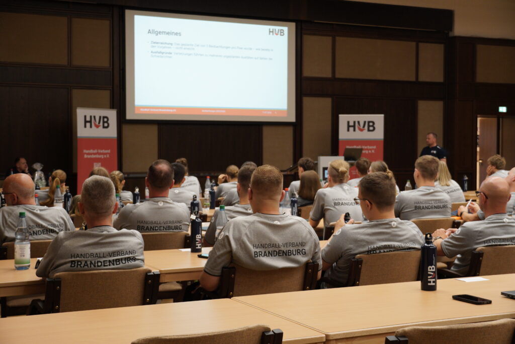 Personen in Trikots des Handball-Verbandes Brandenburg sitzen in einem Konferenzraum und blicken auf einen Vortragenden sowie eine große Leinwand, auf der eine Präsentation gezeigt wird. Auf den Tischen stehen Flaschen und liegen Papiere.