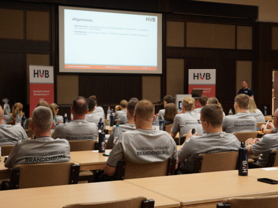 Personen in Trikots des Handball-Verbandes Brandenburg sitzen in einem Konferenzraum und blicken auf einen Vortragenden sowie eine große Leinwand, auf der eine Präsentation gezeigt wird. Auf den Tischen stehen Flaschen und liegen Papiere.