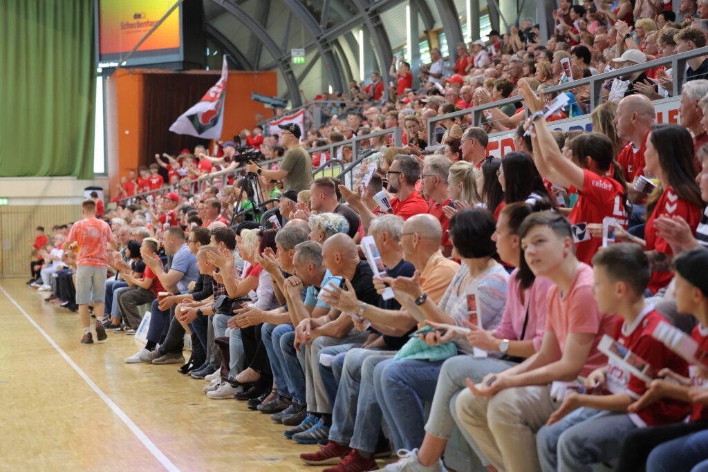 Bei einer Hallensportveranstaltung sitzt eine große Menschenmenge auf den Tribünen; viele sind in Rot und Weiß gekleidet, klatschen und jubeln. Einige halten Fahnen und Transparente hoch, um die Nachwuchsleistungssport-Mannschaften anzufeuern, während andere Fotos oder Videos aufnehmen.