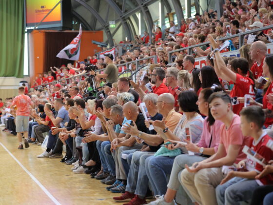 Bei einer Hallensportveranstaltung sitzt eine große Menschenmenge auf den Tribünen; viele sind in Rot und Weiß gekleidet, klatschen und jubeln. Einige halten Fahnen und Transparente hoch, um die Nachwuchsleistungssport-Mannschaften anzufeuern, während andere Fotos oder Videos aufnehmen.