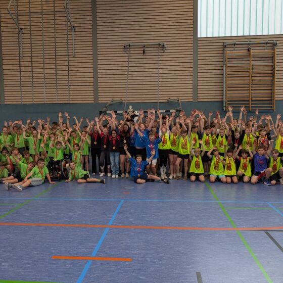Eine große Gruppe von Kindern in Sportuniformen posiert in einer Turnhalle, viele von ihnen heben die Arme. Einige tragen grüne Trikots, andere gelbe. In der Mitte stehen einige Erwachsene, die einen Pokal und Medaillen in den Händen halten.