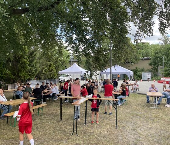 Die Menschen sitzen draußen unter Bäumen an Picknicktischen, im Hintergrund sind weiße Zelte und Imbissstände zu sehen. Im Vordergrund stehen zwei Kinder in roten Sporttrikots neben einem Tisch. Es ist eine entspannte Szene, die ein gemeinschaftliches Beisammensein zeigt.