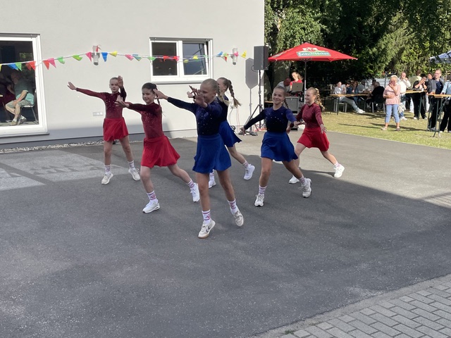 Sechs junge Mädchen in einheitlichen Tanzoutfits führen auf dem Bürgersteig im Freien eine Tanzchoreografie vor, während im Hintergrund Zuschauer zuschauen. Rote und blaue Kostüme sowie bunte Wimpel sorgen für eine festliche Atmosphäre.
