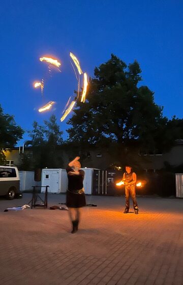 Zwei Menschen führen in der Abenddämmerung im Freien eine Feuerjonglage vor; einer wirft brennende Keulen in die Luft, während der andere Feuerrequisiten hält, im Hintergrund sind Bäume, ein Transporter und Gebäude zu sehen.