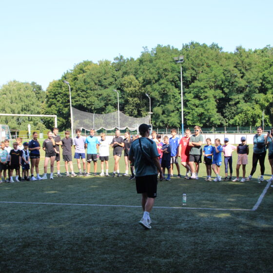 Eine Gruppe von Menschen steht in einem großen Kreis auf einem grasbewachsenen Sportplatz und hört einer Person zu, die in der Mitte spricht. Im Hintergrund sind Bäume und Sportgeräte zu sehen.