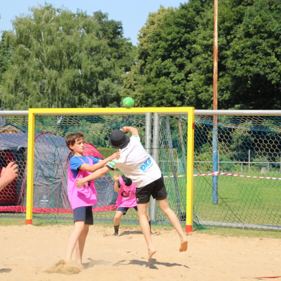 Kinder spielen Strandhandball; ein Kind in Weiß springt hoch, um einen grünen Ball aufs Tor zu werfen, während ein anderes Kind in Rosa versucht, den Wurf zu blocken. Im Hintergrund sind Bäume, Gras und ein Zaun zu sehen.