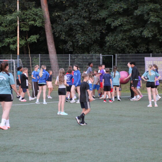 Eine Gruppe von Kindern und Jugendlichen spielt und tobt auf einem Sportplatz im Freien, der von Bäumen und einem Metallzaun umgeben ist, während einige Erwachsene auf sie aufpassen.