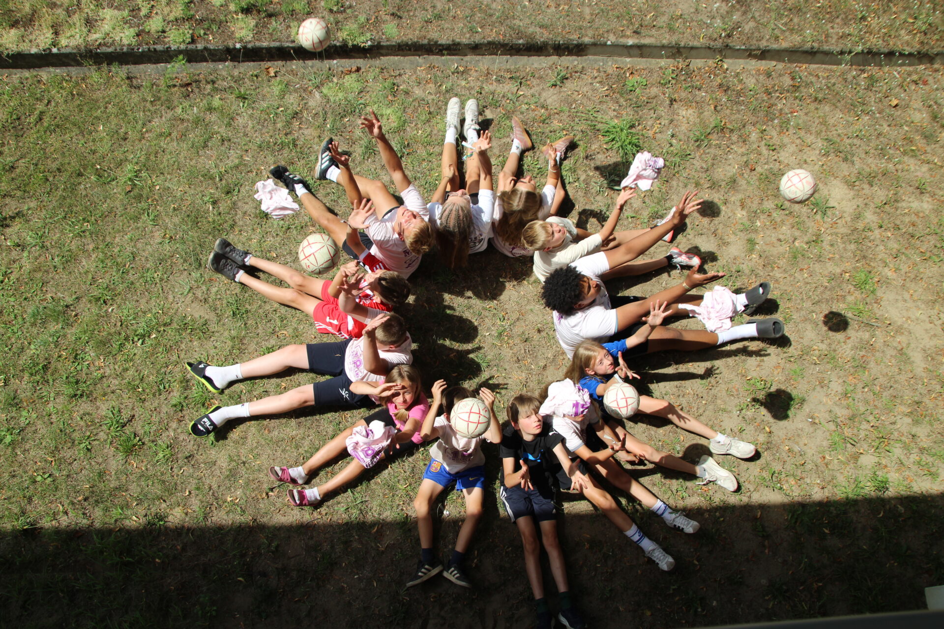 Eine Gruppe von Kindern liegt im Kreis auf dem Rasen, die Füße berühren sich in der Mitte, während sie Volleyballbälle in die Luft werfen und so ein verspieltes, symmetrisches Muster bilden.