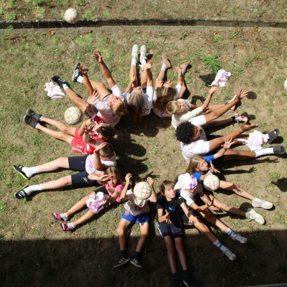 Eine Gruppe von Kindern liegt im Kreis auf dem Rasen, die Füße berühren sich in der Mitte, während sie Volleyballbälle in die Luft werfen und so ein verspieltes, symmetrisches Muster bilden.