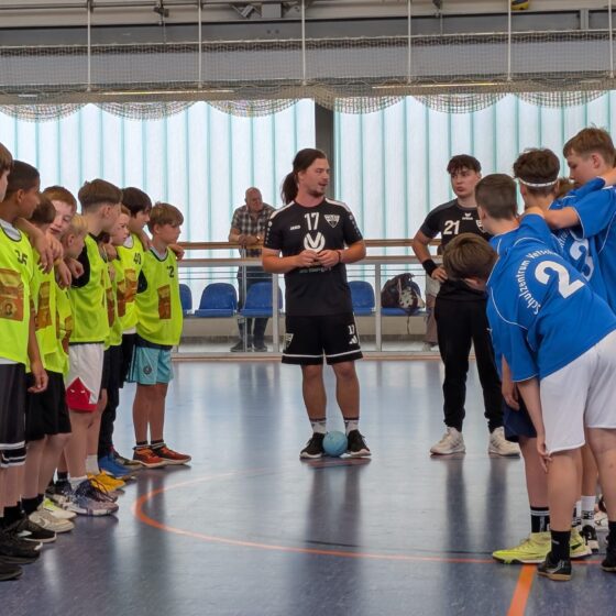 Zwei Jugendhandballmannschaften – die eine in grünen Überziehhemden, die andere in blauen Trikots – stehen sich auf einem Hallenplatz gegenüber, während zwei Trainer zwischen ihnen stehen und sich auf ein Spiel vorbereiten.