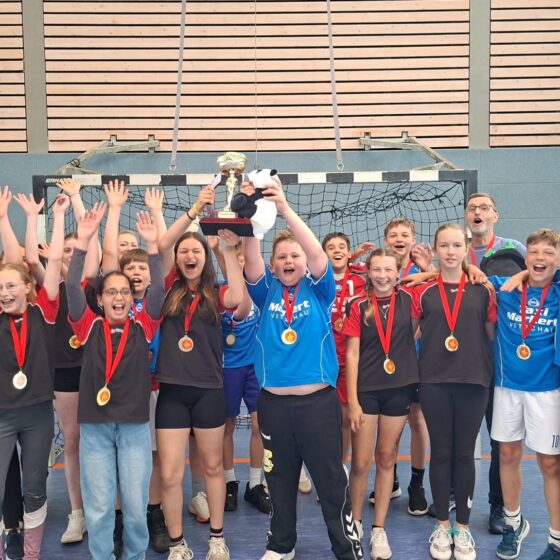 Eine Gruppe von Kindern mit Medaillen jubelt und reckt die Hände in die Höhe, während sie in einer Sporthalle einen Pokal hochhalten und einen sportlichen Sieg feiern.