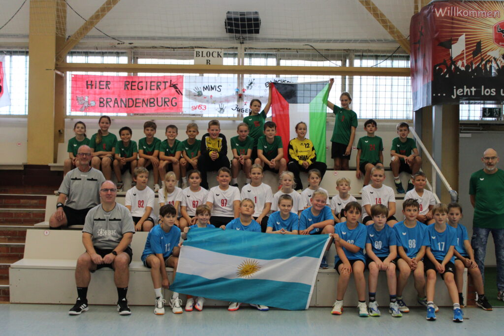 Eine große Gruppe junger Handballspieler und -trainer posiert beim HVB-Pokal-Final-Four 2026/27 auf den Tribünen der Halle mit palästinensischen und argentinischen Flaggen.